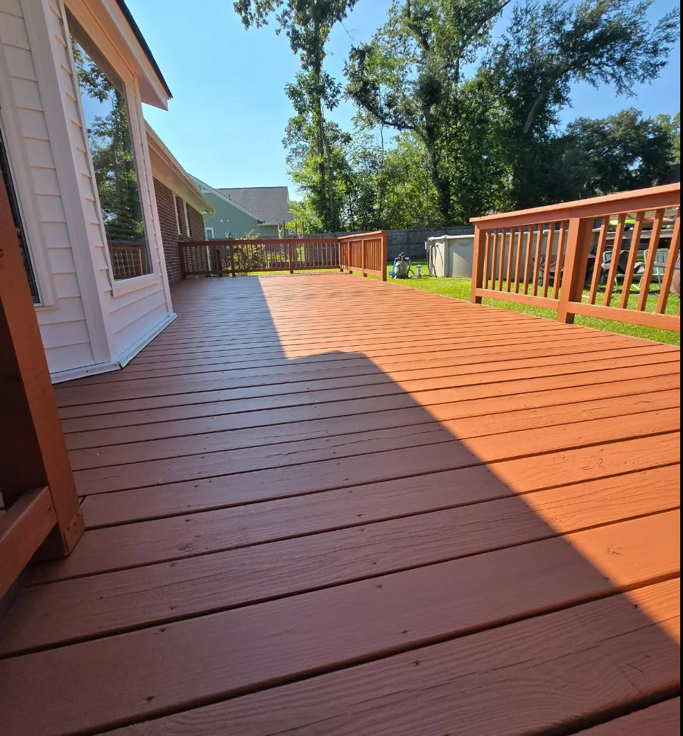 Backyard wood deck and railings finished in a warm redwood-toned solid stain beside a white lap-sided house, by Margarito's Service Painting