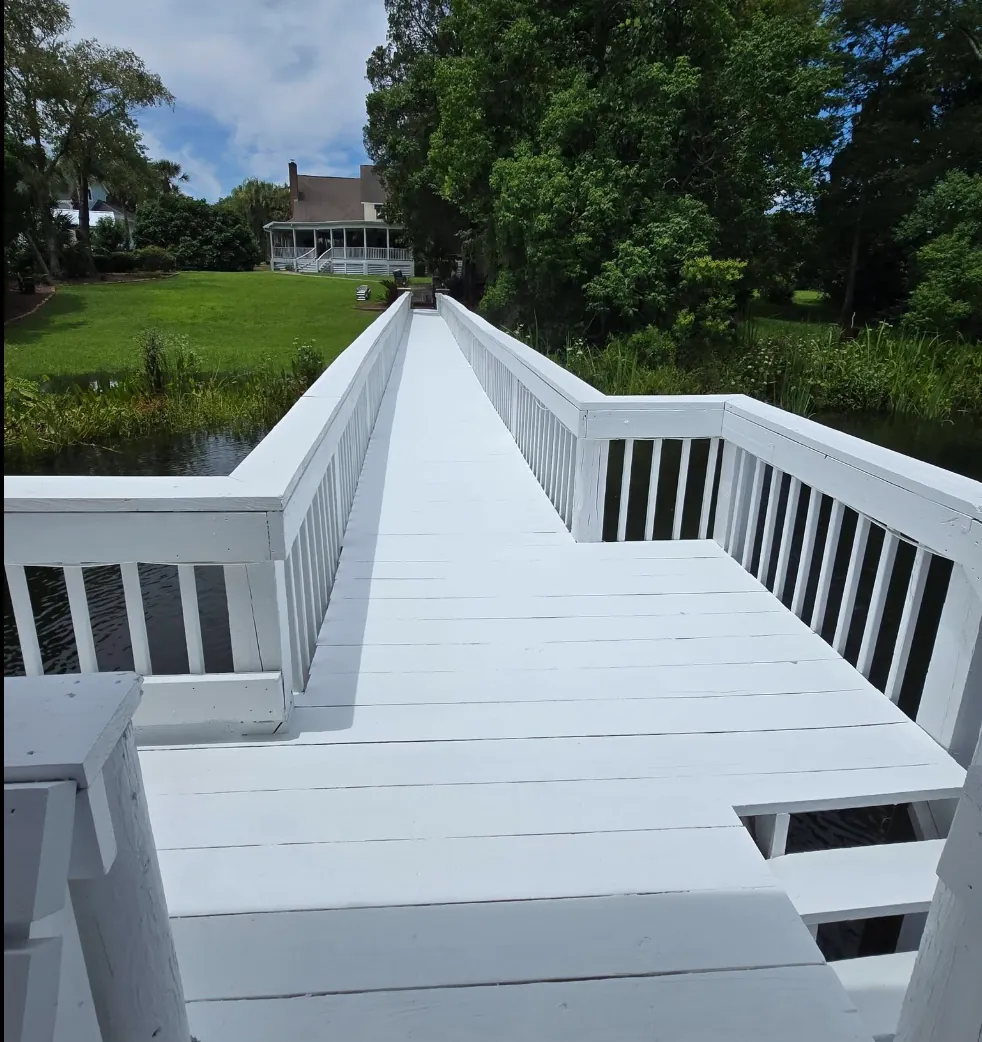 Freshly painted bright white wooden dock and walkway with railings crossing a pond toward a home with a screened porch, by Margarito's Service Painting