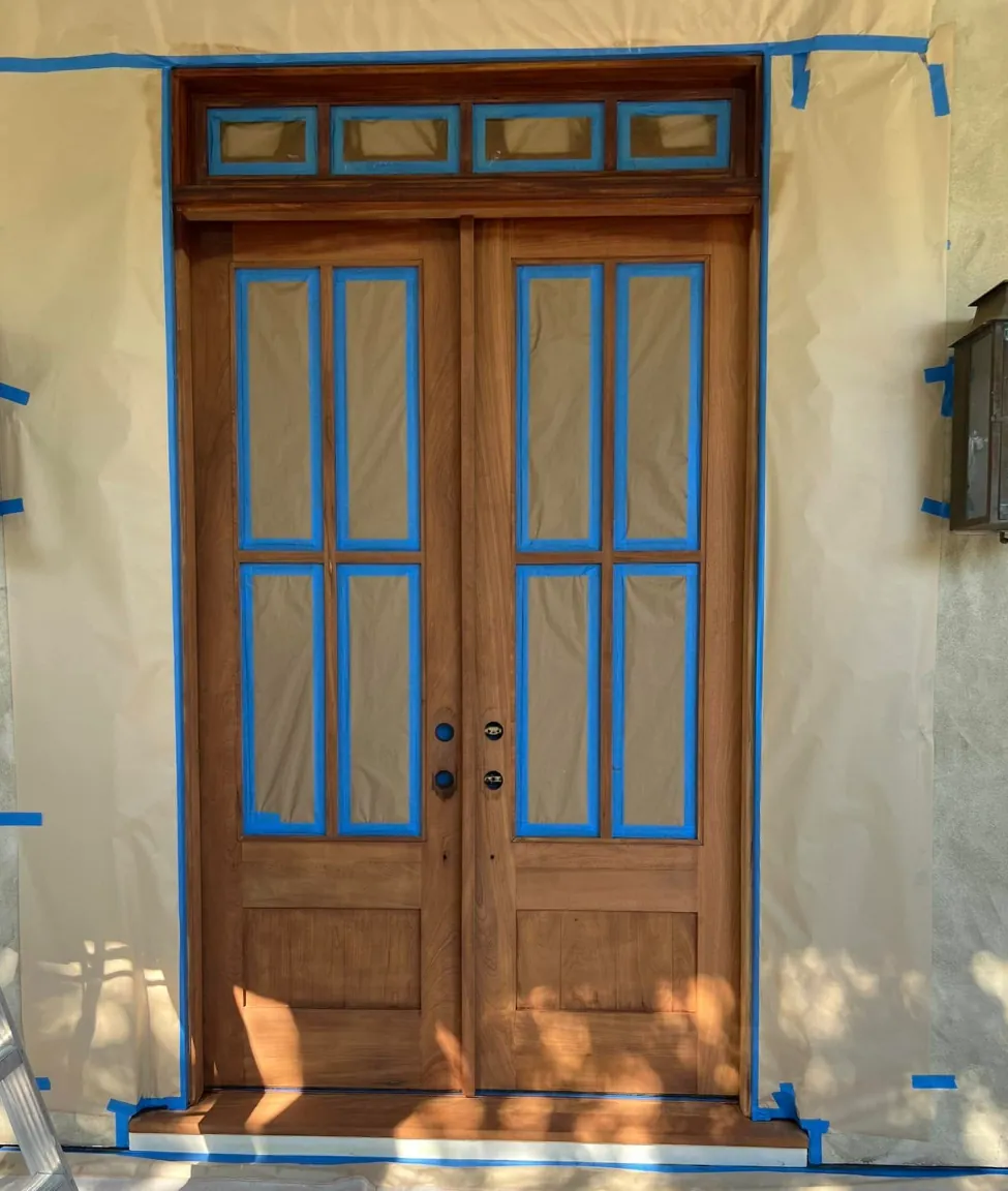 Stained wood double entry door with glass panes and transom fully masked in blue tape and paper, surrounding stucco wall papered off, by Margarito's Service Painting