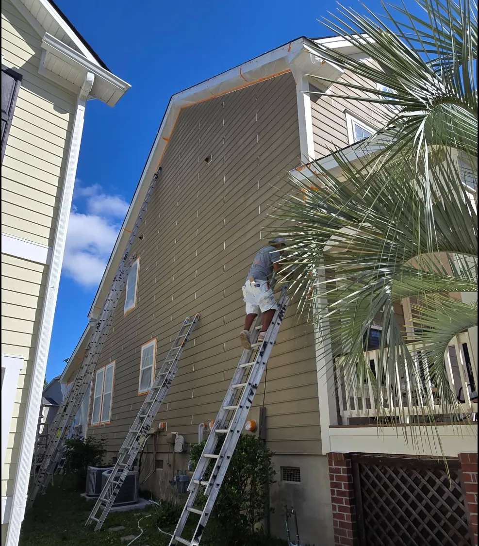 Painter from Margarito's Service Painting on an extension ladder coating the tan lap siding of a two-story Lowcountry home, palmetto fronds in the foreground